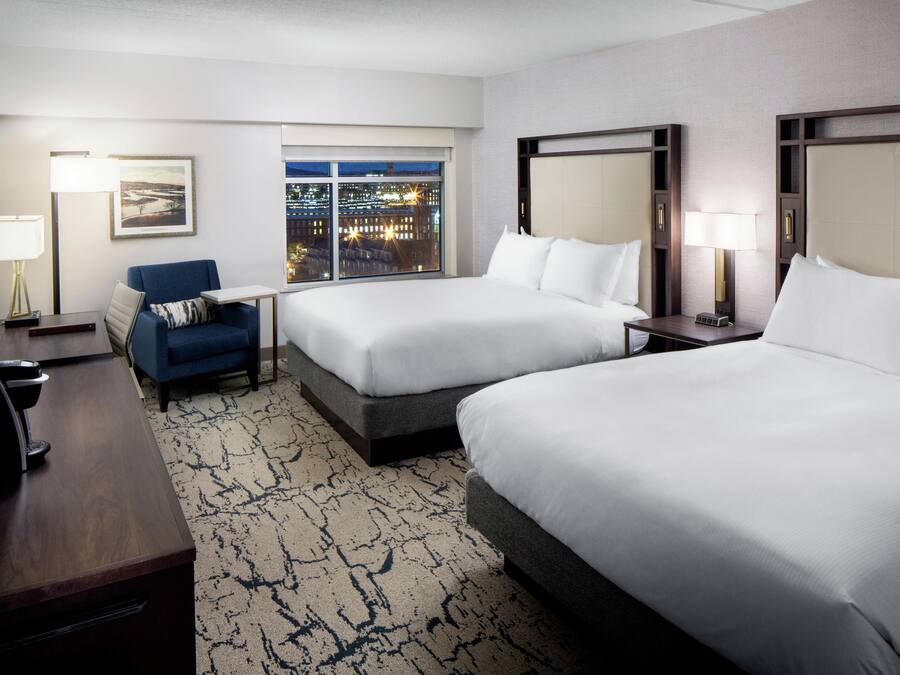 A hotel room with two beds, a desk, a chair, a TV, and a window with a city view.