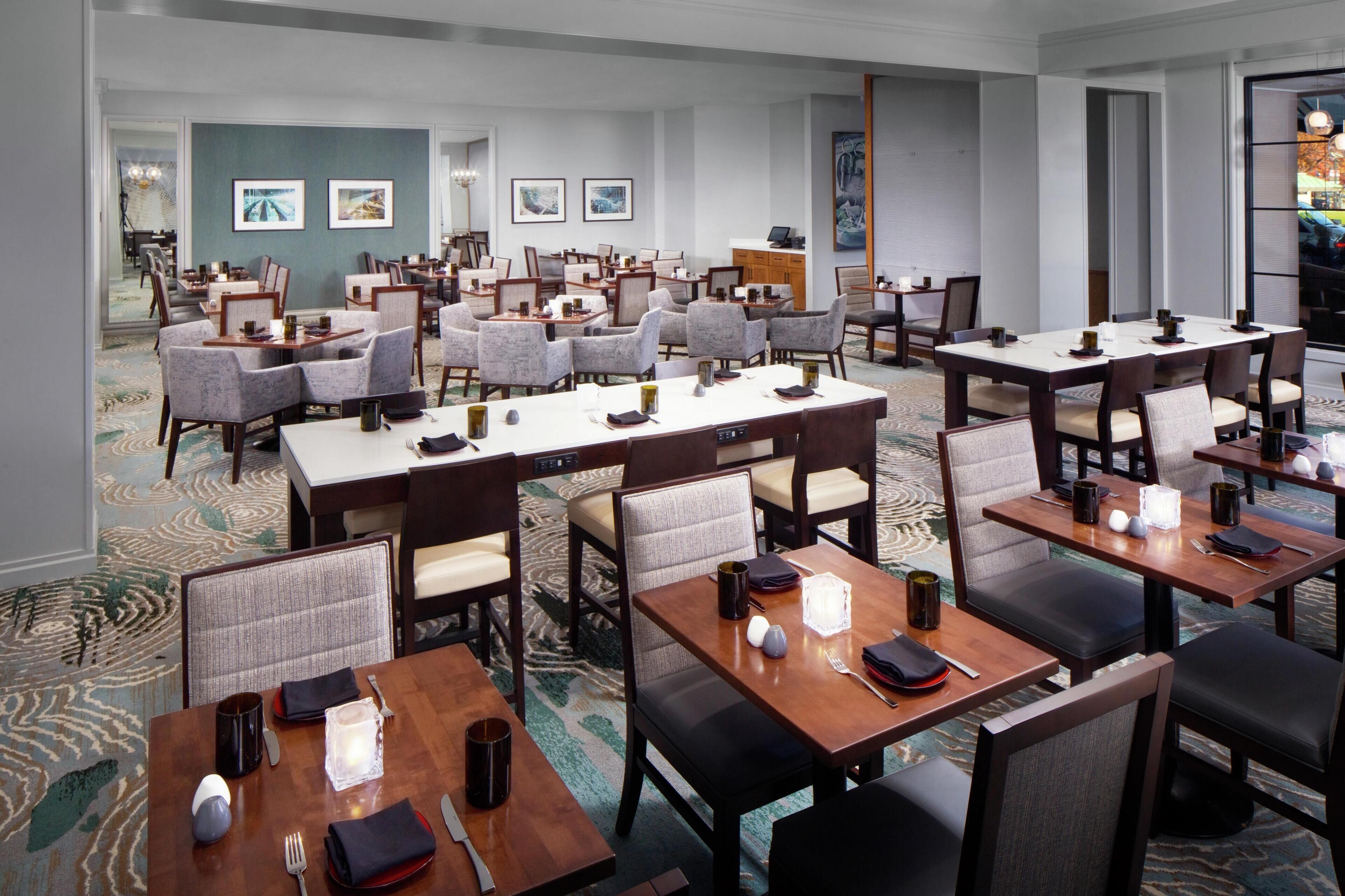 A hotel dining area with multiple tables and chairs, a carpeted floor, and framed artwork on the walls.