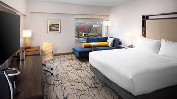 A hotel room with a large bed, a desk with a computer, a chair, a sofa, and a window with a view of trees and buildings.