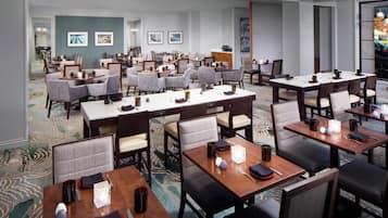 A hotel dining area with multiple tables and chairs, a carpeted floor, and framed artwork on the walls.
