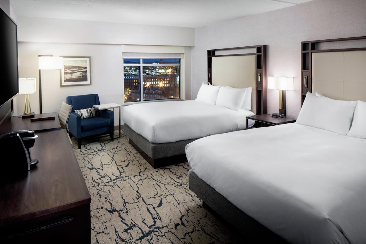 Quarto de hotel com duas camas, uma escrivaninha, uma cadeira, uma TV e uma janela com vista para a cidade.