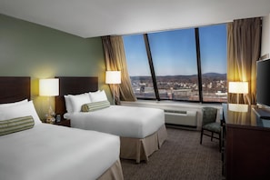 A hotel room with two beds, a desk, a chair, a TV, and a large window with a city view.