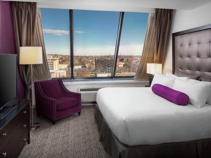 Una habitación de hotel con un ventanal grande, una cama, una silla, un televisor y un aparador.