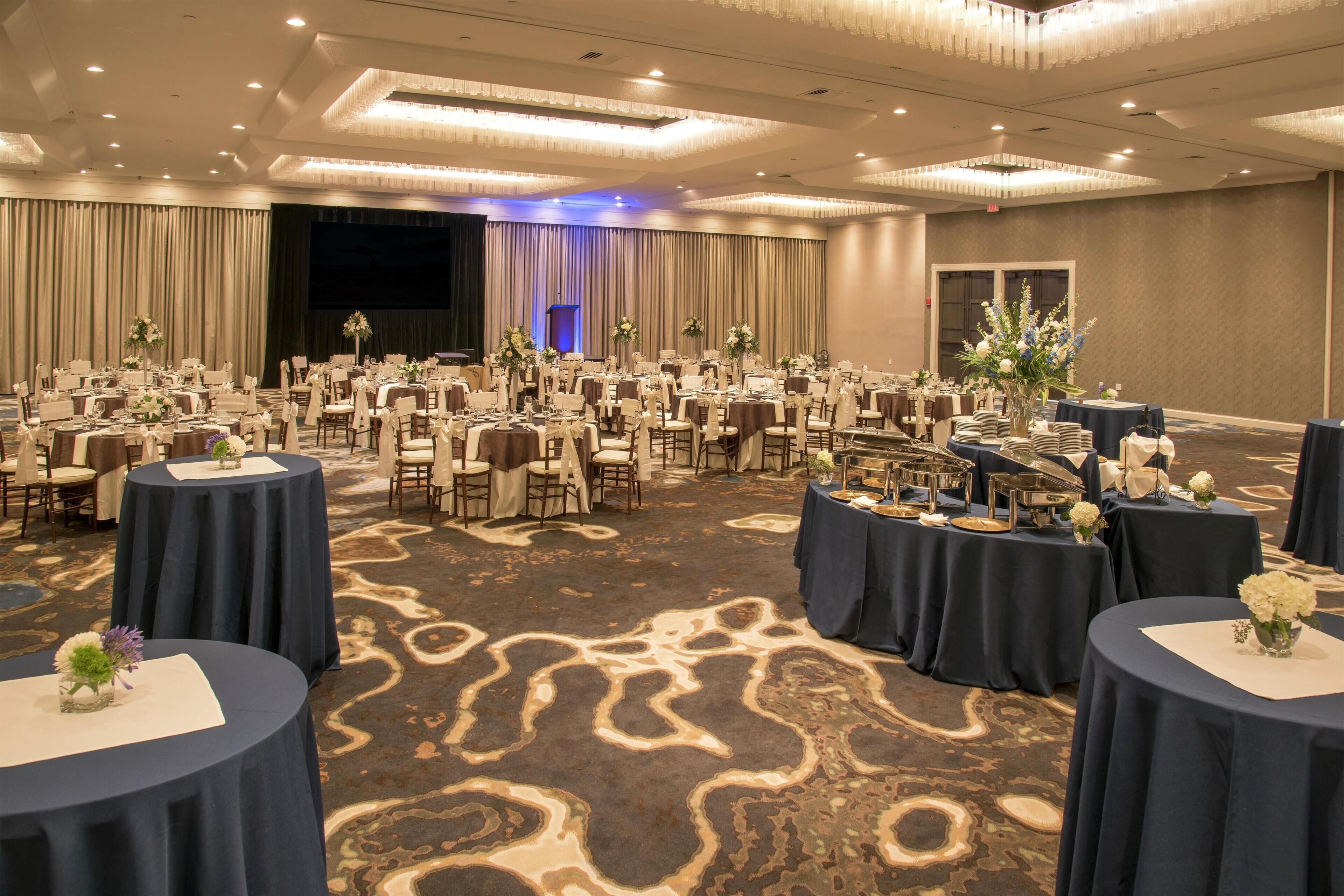 Um amplo salão de banquetes com mesas redondas, cadeiras e centros de mesa florais.