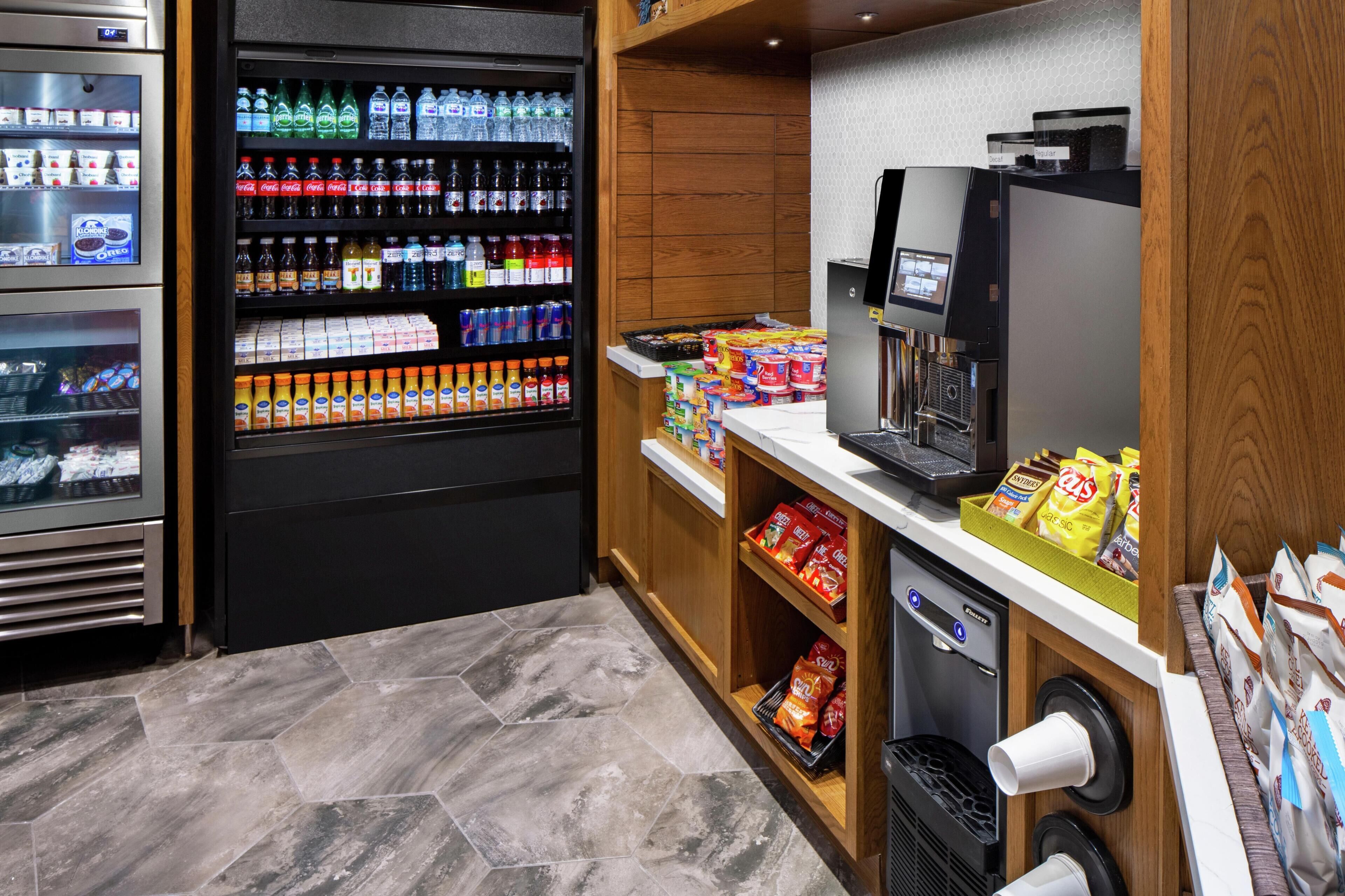 A self-service beverage kiosk with refrigerated drinks and snacks, a coffee machine, and a trash bin.