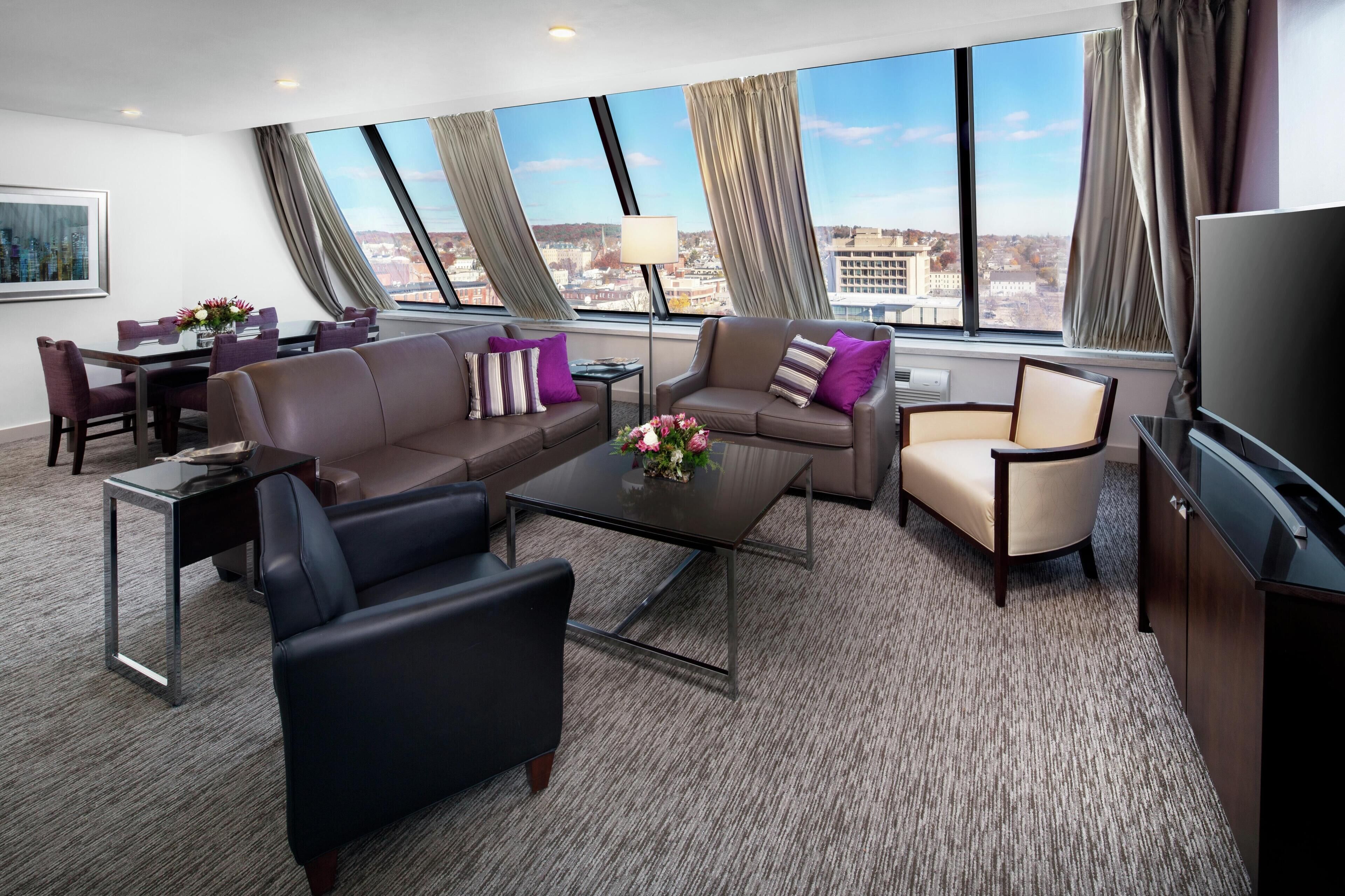 A modern hotel room with a large window, a sofa set, a coffee table, a TV, and a dining area.