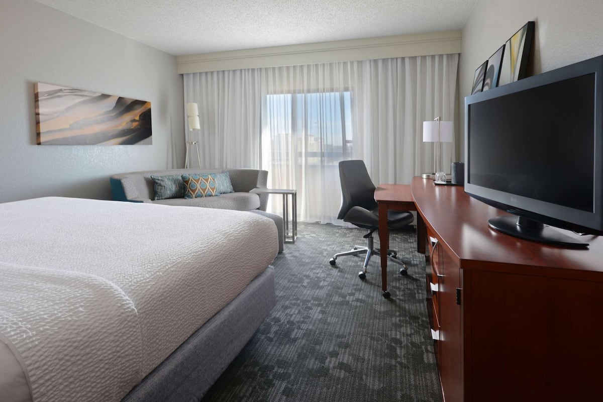 A hotel room with a large bed, a desk with a flat-screen TV, a chair, a small table, and a window with curtains.