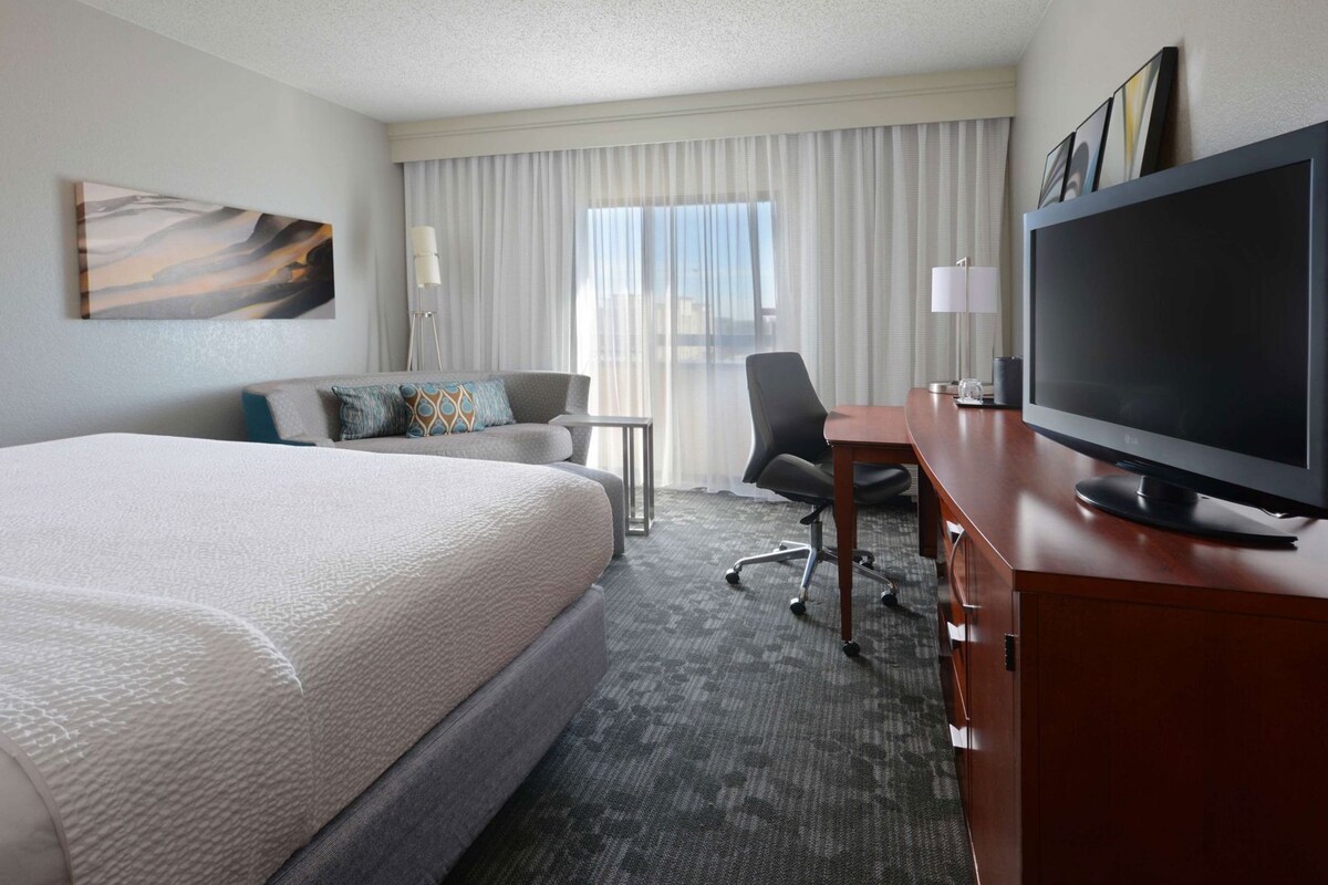 A hotel room with a large bed, a desk with a flat-screen TV, a chair, a small table, and a window with curtains.