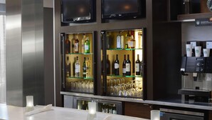 Uma área de bar com prateleiras cheias de garrafas de bebidas e copos, uma máquina de café e uma televisão de tela plana fixada na parede.