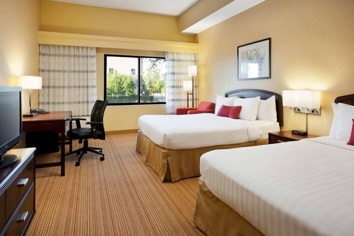 A hotel room with two beds, a desk with a chair, a television, and a window with curtains.