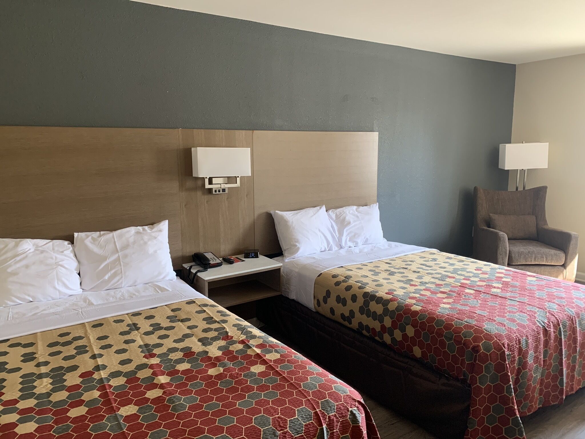 A hotel room with two beds, a nightstand, a chair, and a wall-mounted lamp.