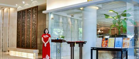 A modern interior with a woman in a red dress standing near a glass wall, a wooden panel, and a table with framed artwork.