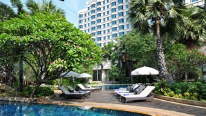 A pool area with lounge chairs and umbrellas, surrounded by lush greenery and a tall building in the background. - Rama Gardens Hotel Bangkok (Bangkok)
