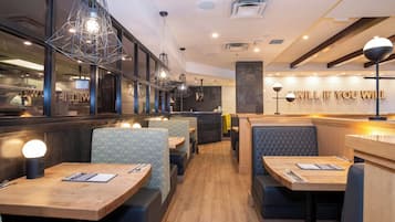 A restaurant interior with wooden tables, cushioned seating, pendant lighting, and a brick wall with illuminated text.