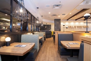 A restaurant interior with wooden tables, cushioned seating, pendant lighting, and a brick wall with illuminated text.