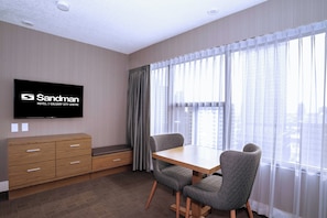 Une chambre d’hôtel avec un meuble en bois, une télévision, une table à manger avec deux chaises et une grande fenêtre ornée de rideaux légers.