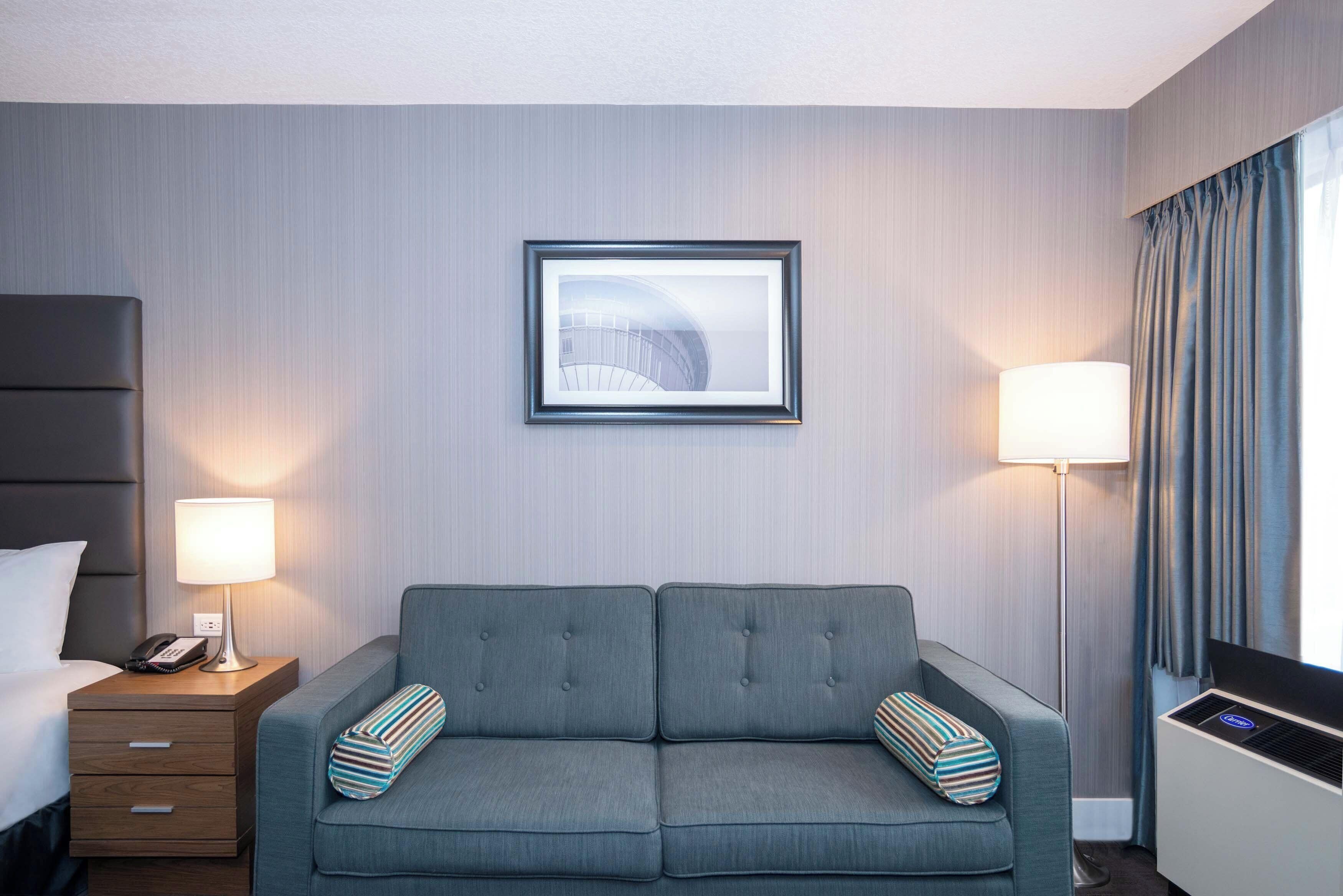 Habitación de hotel con un sofá gris, una mesita de noche con una lámpara, un cuadro en la pared y una ventana con cortinas.