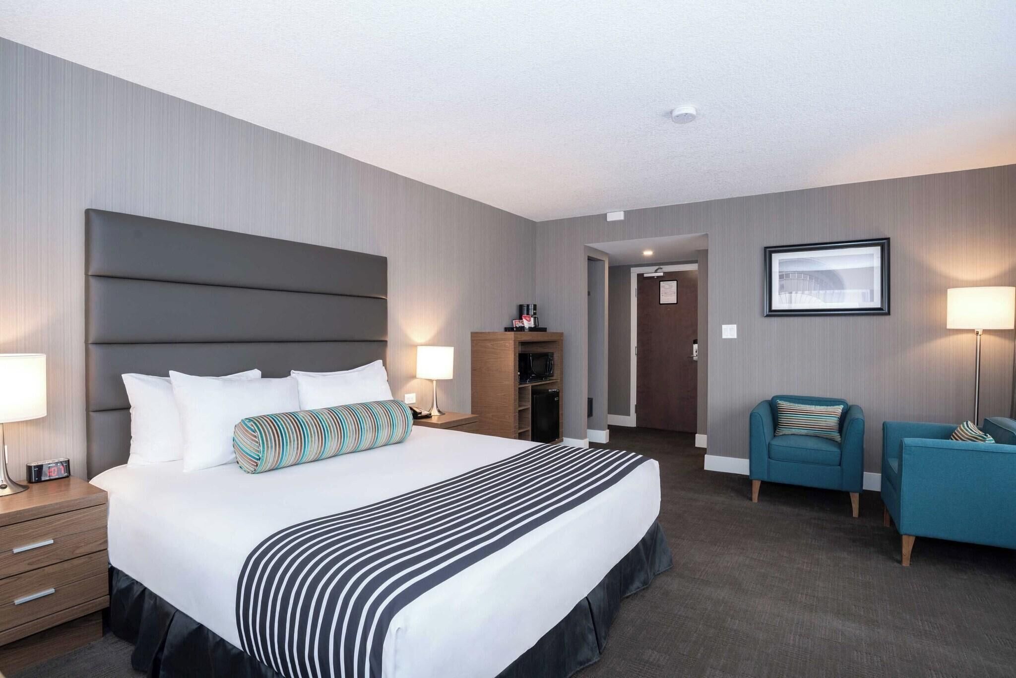 Habitación de hotel con una cama grande, mesitas de noche, un armario, una zona de estar con una silla y un sofá, y un cuadro enmarcado en la pared.