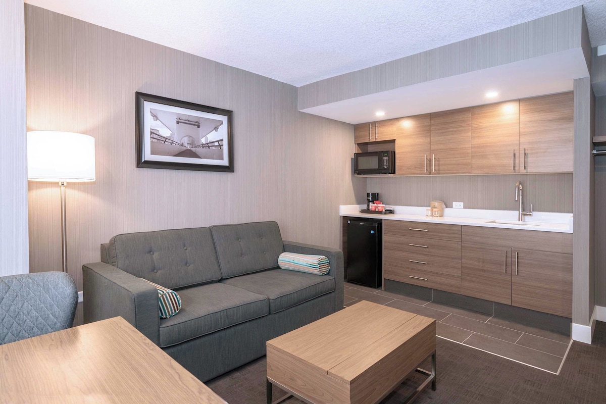 A modern kitchen with a microwave, sink, and wooden cabinets. There is a grey sofa with a striped pillow and a lamp on the left.