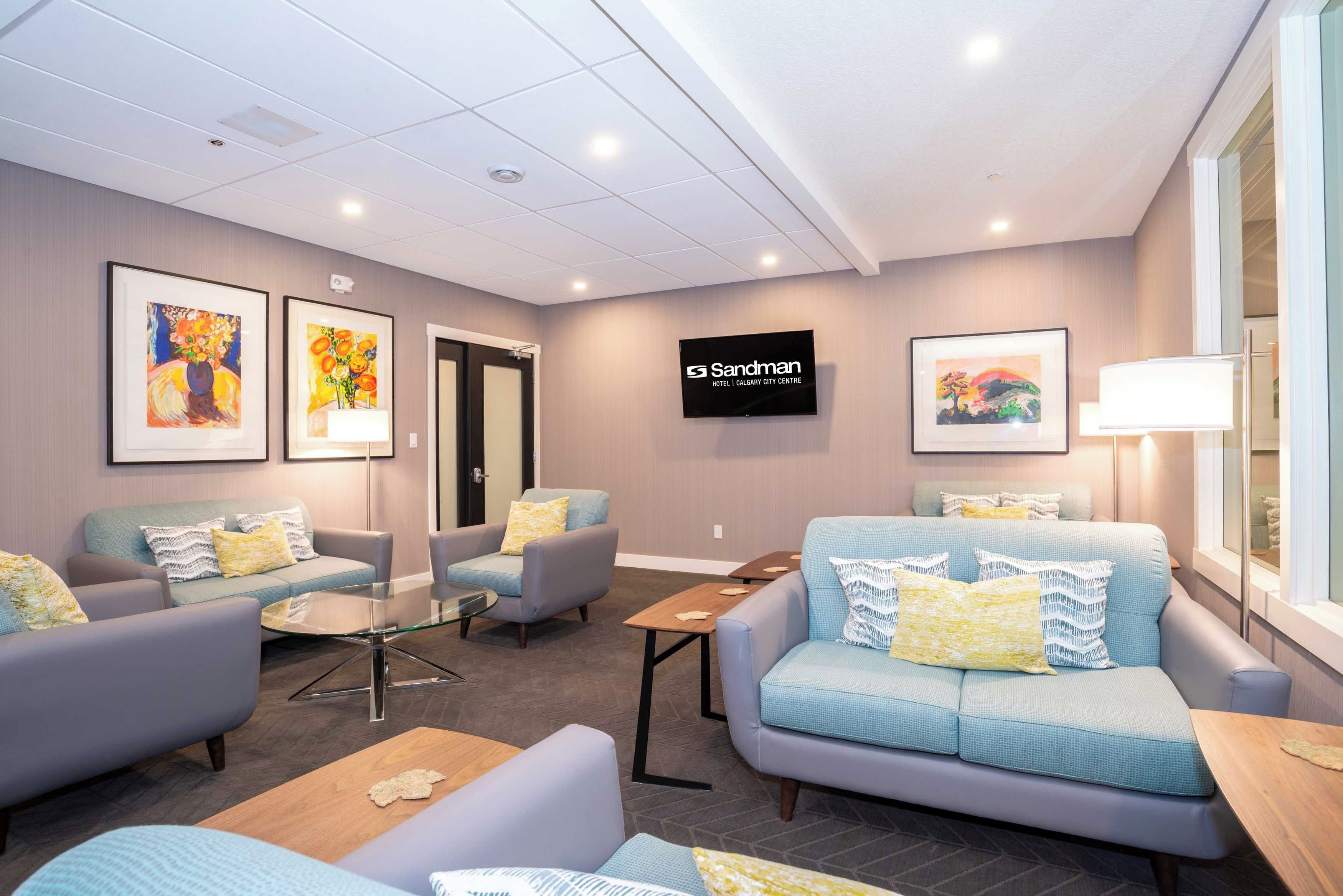 Eine Hotelhalle mit Sitzbereichen, Kunstwerken an den Wänden, einem Fernseher und einem Schild für das Sandman Hotel Calgary City Centre.