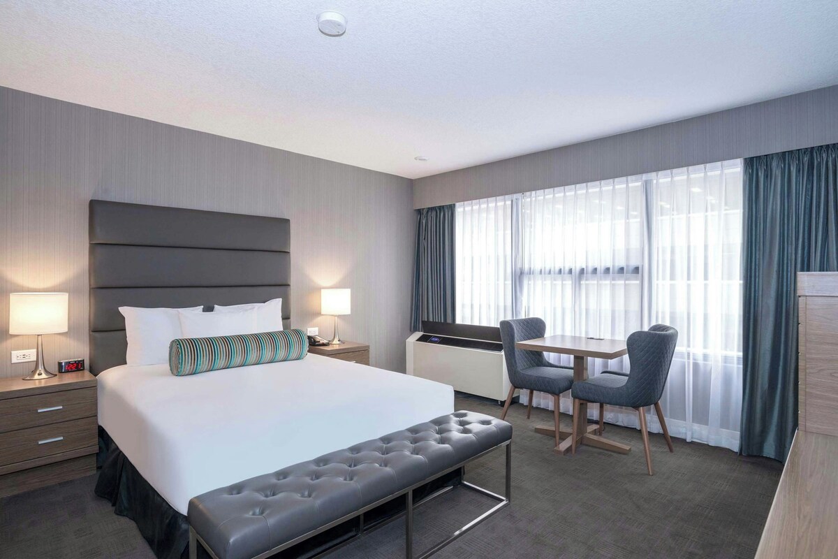 A hotel room with a bed, bedside tables, a desk with a chair, and a window with curtains.