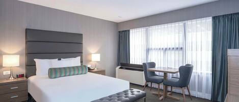 A hotel room with a bed, bedside tables, a desk with a chair, and a window with curtains.