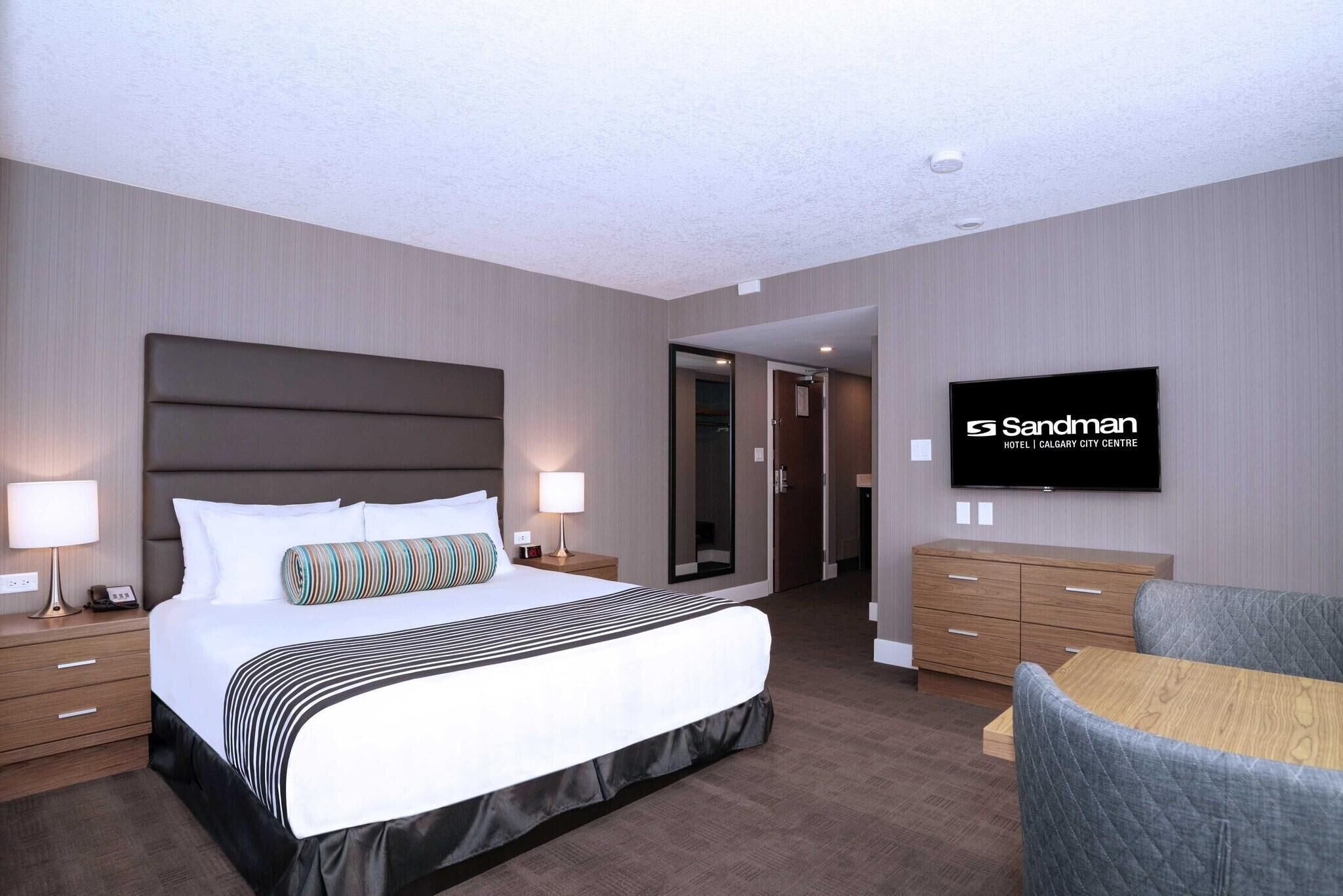 Habitación de hotel con una cama grande, mesitas de noche, televisor, cómoda de madera y un espacio de descanso.