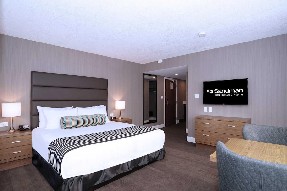 A hotel room with a large bed, bedside tables, a TV, a wooden dresser, and a seating area.
