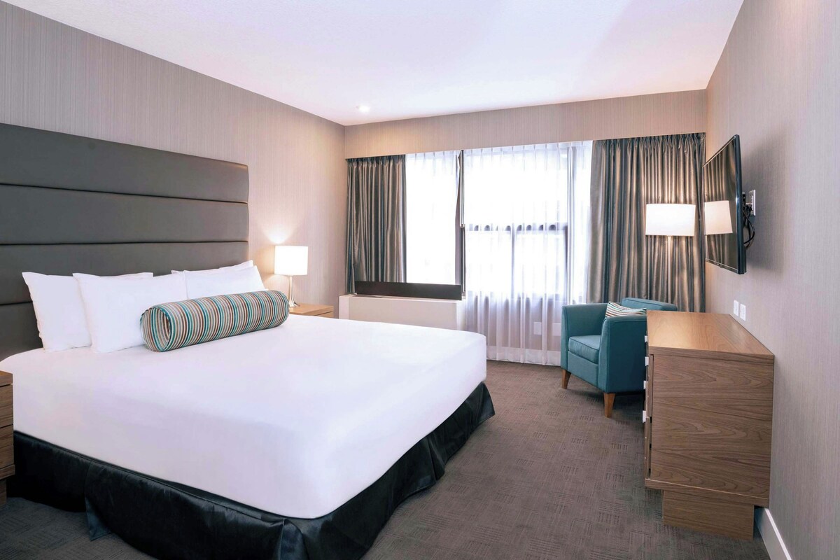 A hotel room with a large bed, a desk, a chair, a TV, and a window with curtains.