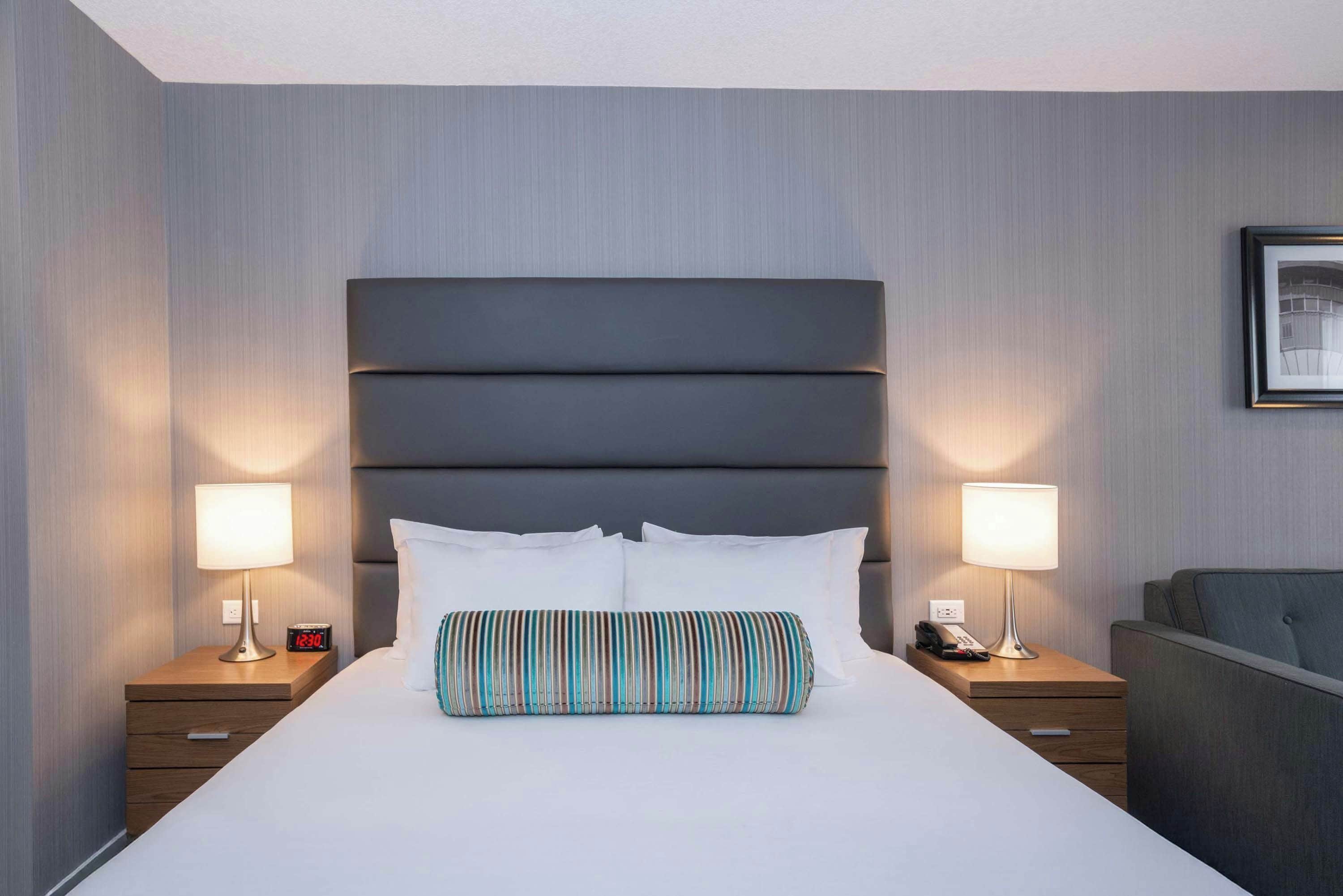 Quarto de hotel com uma cama grande, duas mesas de cabeceira, um criado-mudo, telefone e uma fotografia emoldurada na parede.