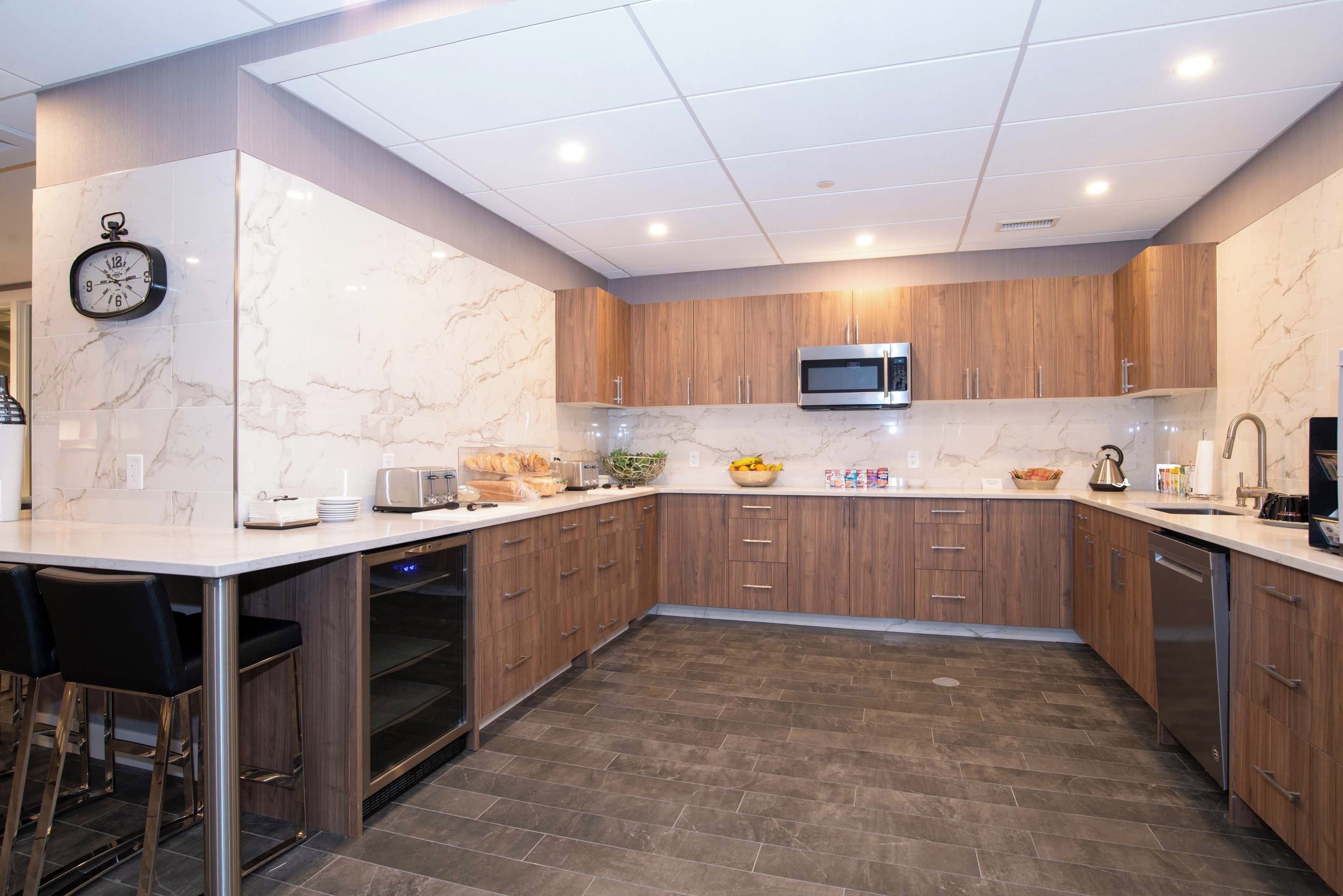A modern kitchen with marble countertops, wooden cabinets, a built-in microwave, and a clock on the wall.