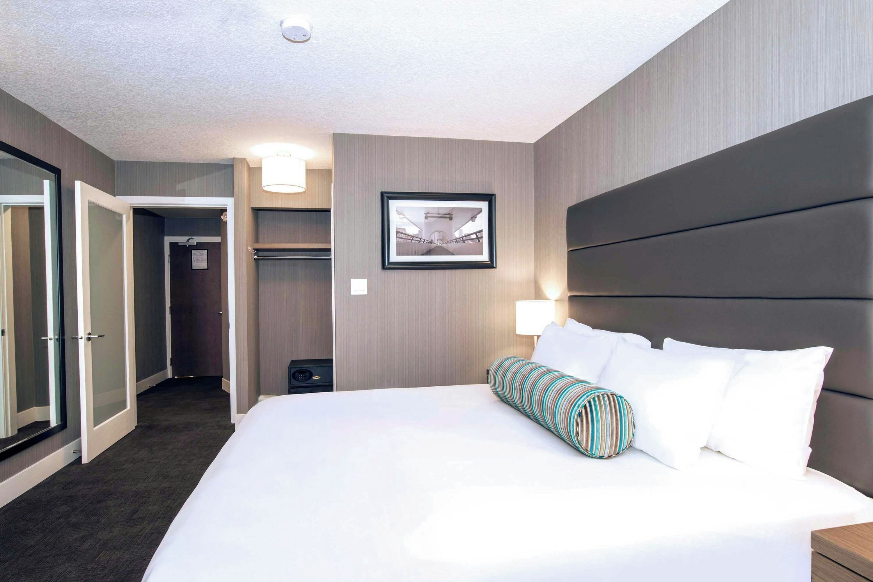 Habitación de hotel con una cama grande, cabecera, mesita de noche, lámpara, un cuadro enmarcado en la pared y una puerta que da a un armario.