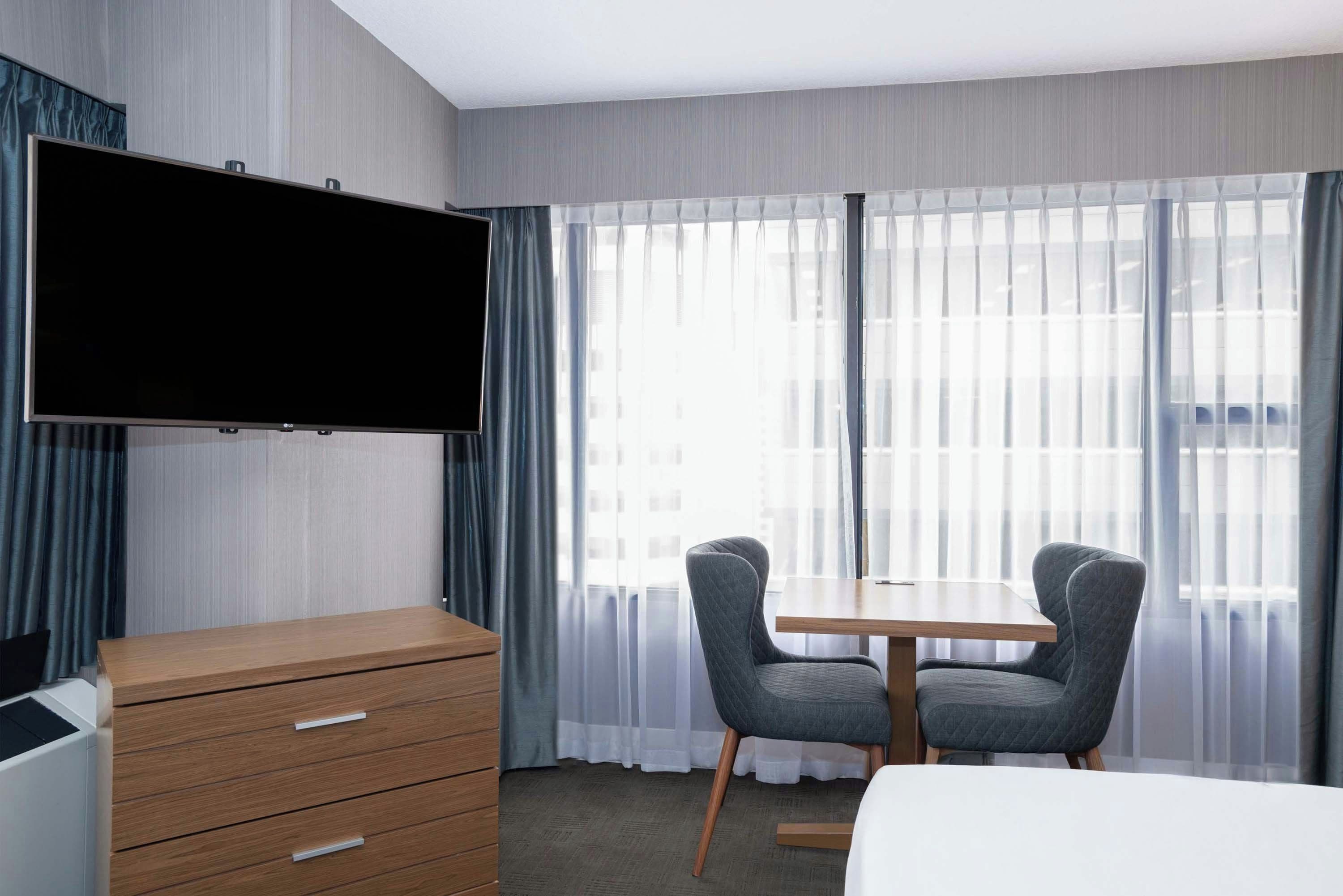 Et hotelværelse med et fladskærms-TV på væggen, en trækommode med skuffer, to polstrede stole omkring et træbord, og et stort vindue med lette gardiner.