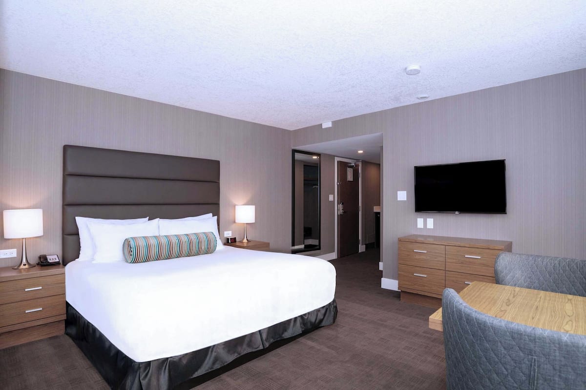Habitación de hotel con una cama grande, mesitas de noche con lámparas, un televisor de pantalla plana en la pared, un aparador de madera y una pequeña mesa con una silla.