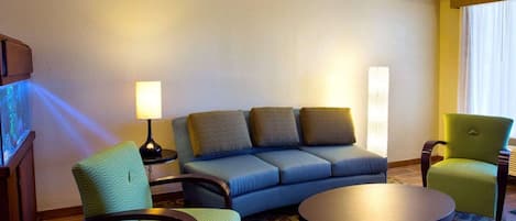 Habitación de hotel con sofá azul, sillón verde, una mesa de centro redonda y una lámpara.