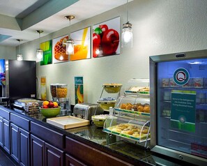 A breakfast buffet with various food items, a refrigerator with a sign indicating items are available only during breakfast hours, and a wall with fruit and vegetable images.