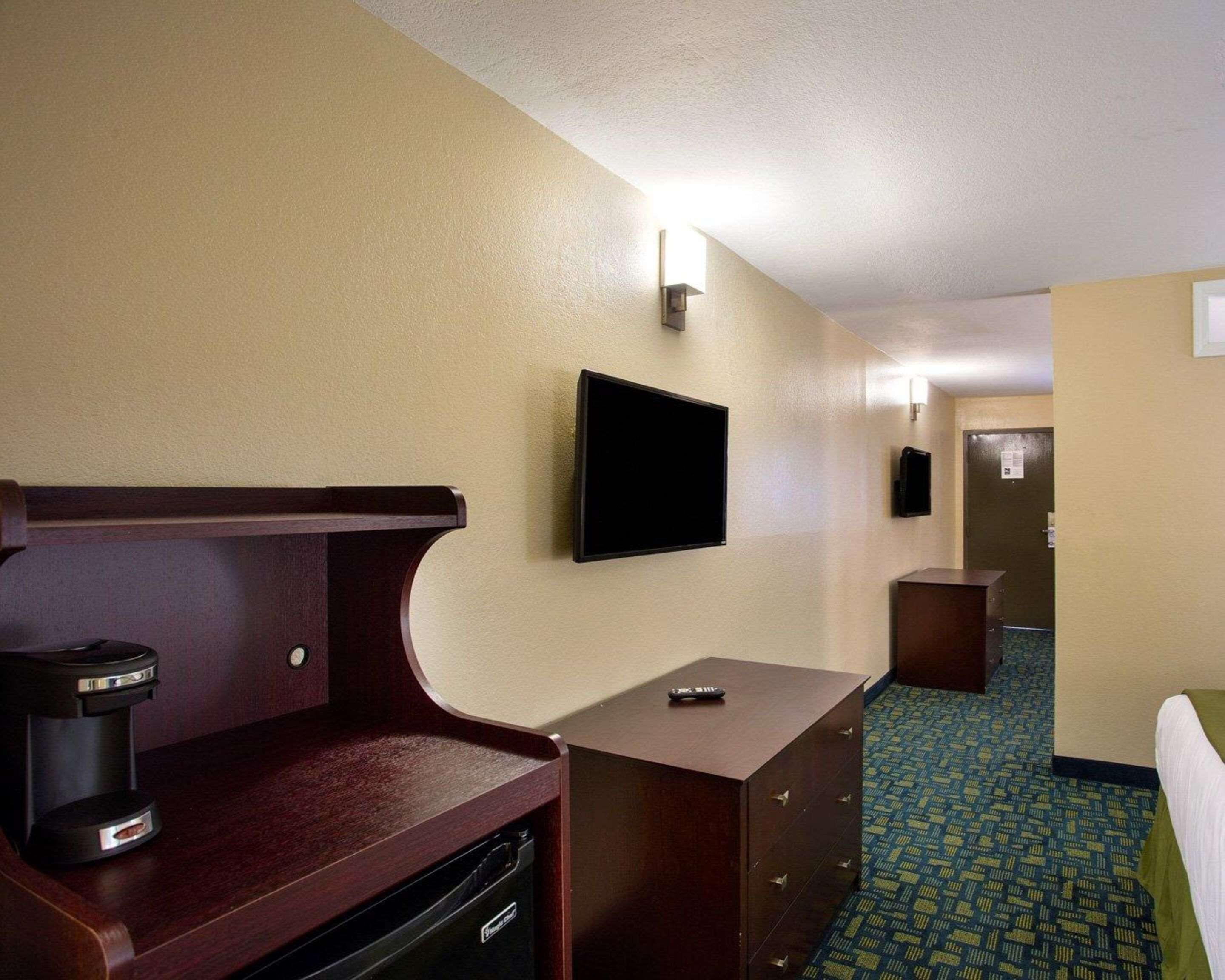 Habitación de hotel con cama, escritorio con cafetera, televisor montado en la pared y alfombra estampada.
