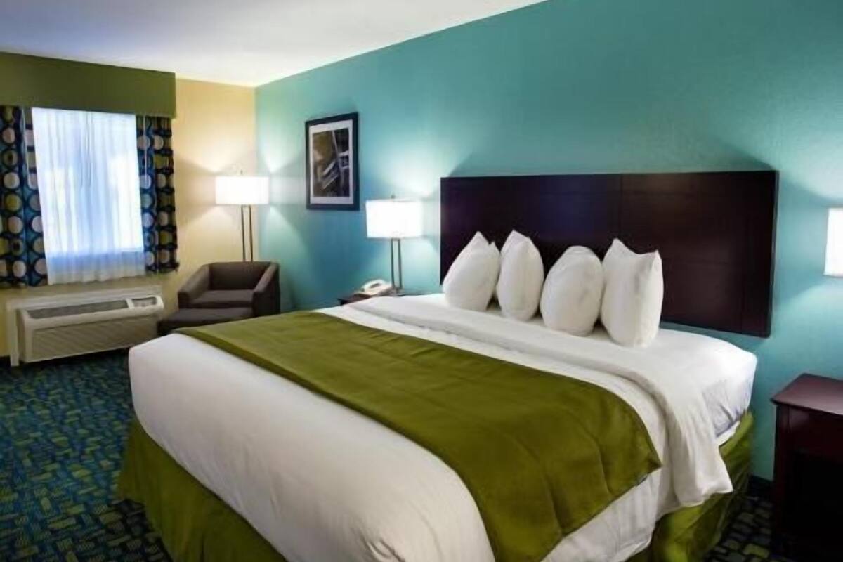 A hotel room with a bed, two bedside lamps, a chair, a window with curtains, and a wall-mounted phone.