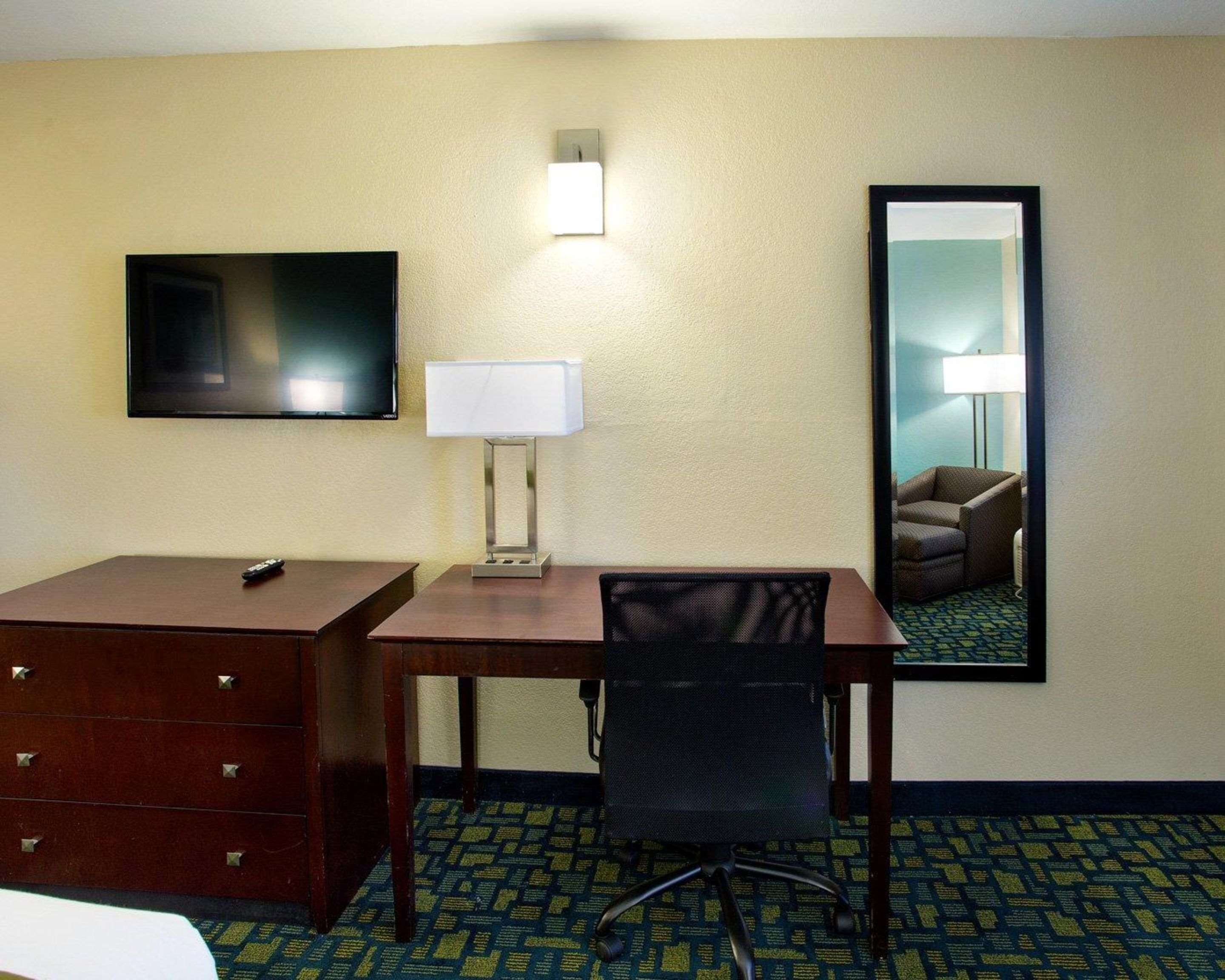 A hotel room with a desk, chair, TV, lamp, dresser, and a mirror.
