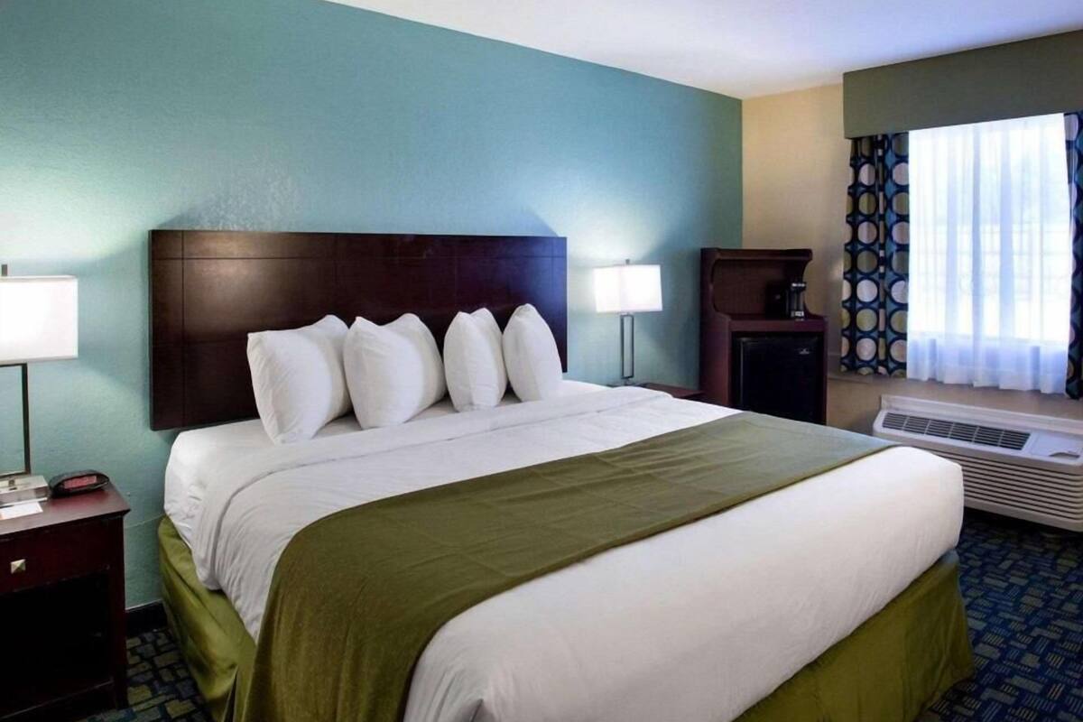 A hotel room with a large bed, a bedside table with a lamp, a small desk with a lamp, a heater, and a window with curtains.