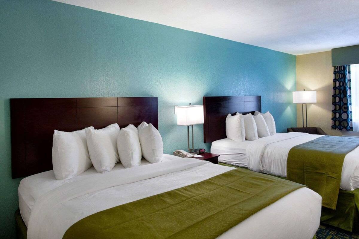 A hotel room with two beds, each with a dark headboard and white bedding. There are two bedside tables with lamps, and the walls are painted in a teal color.