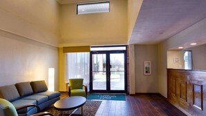 A hotel lobby with a sofa, chairs, a small table, a reception desk, and a large glass door leading outside.
