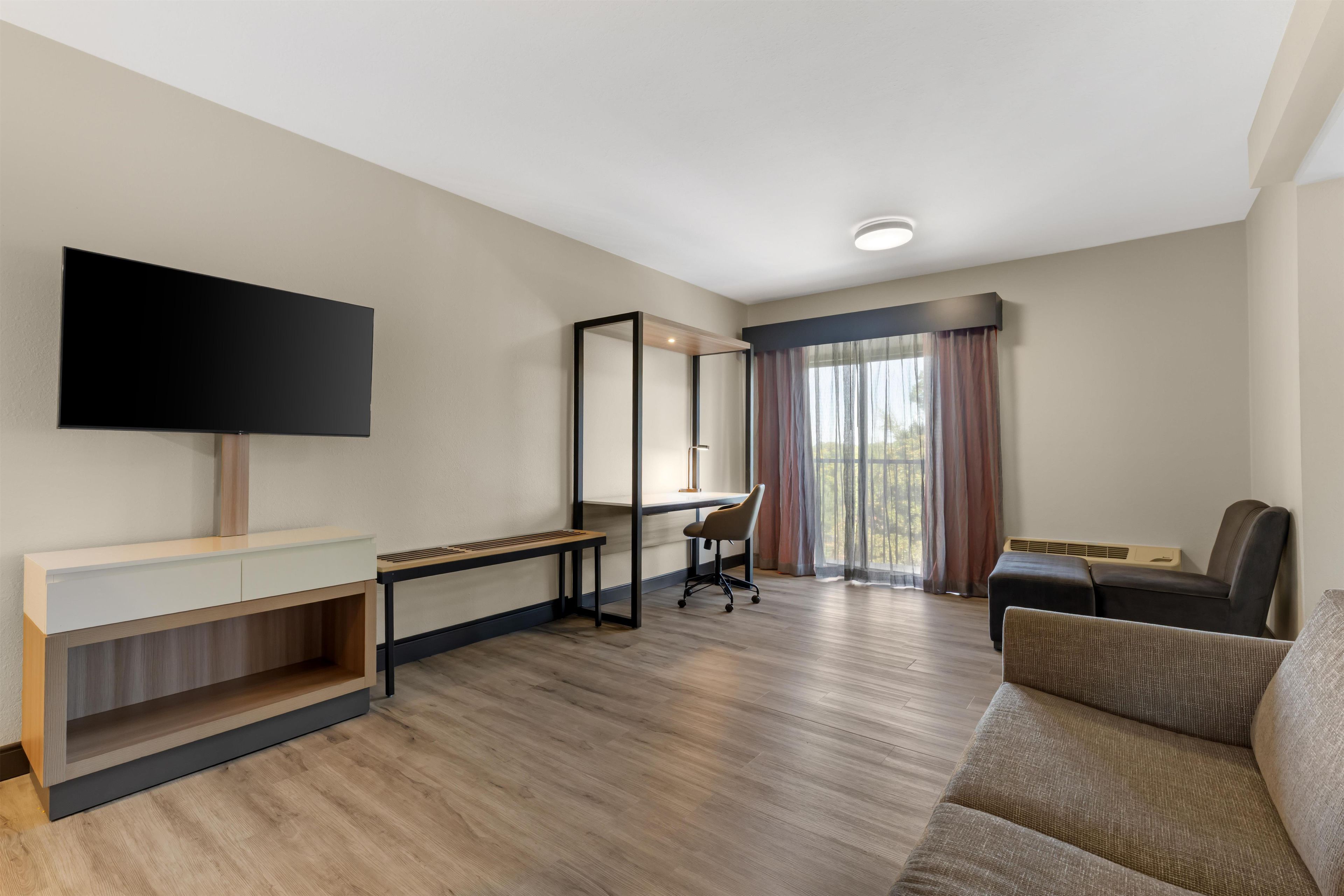 A modern hotel room with a flat-screen TV, a desk with a chair, a large window with curtains, and a sofa.