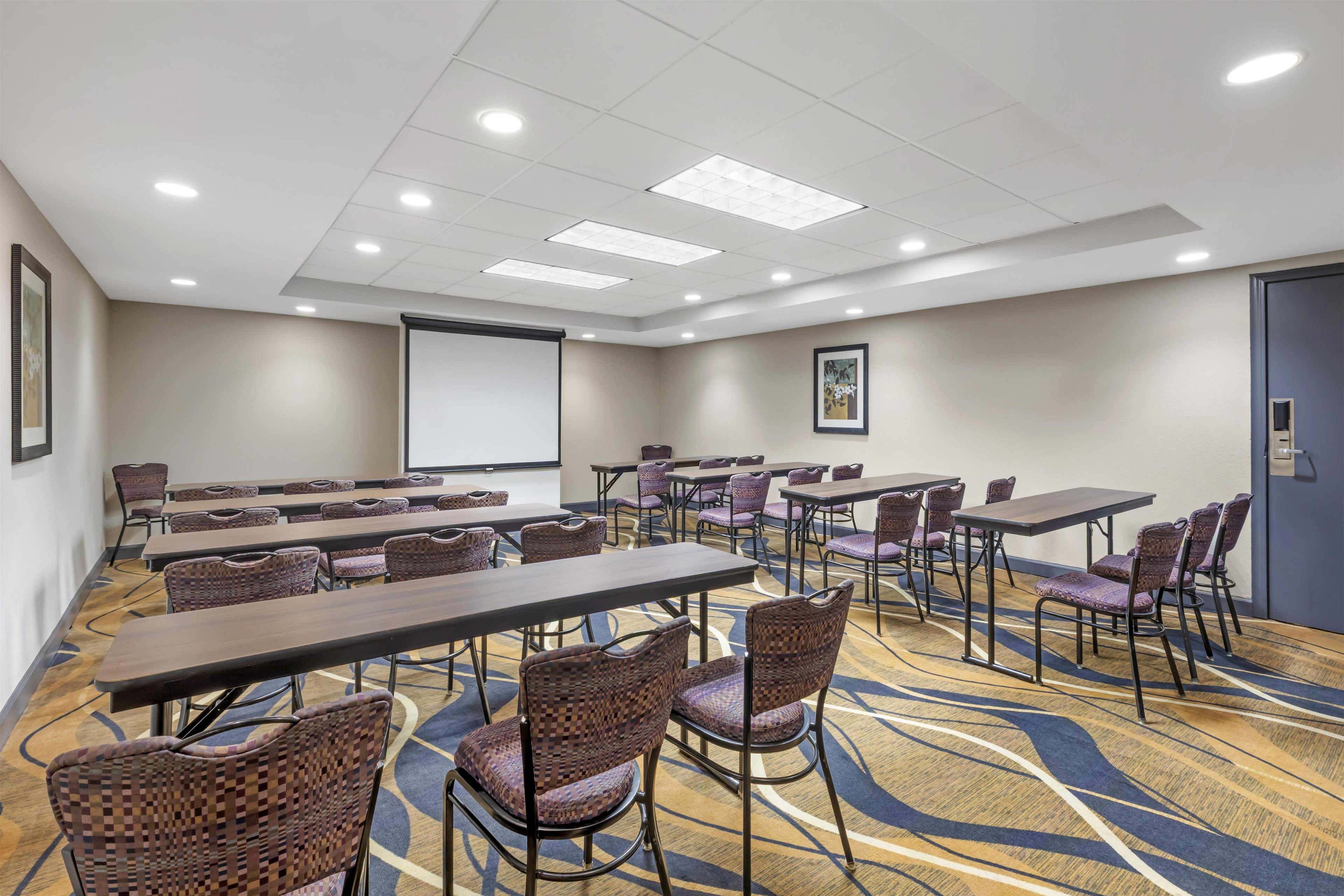 A conference room with multiple tables and chairs, a projector screen, and a carpeted floor.