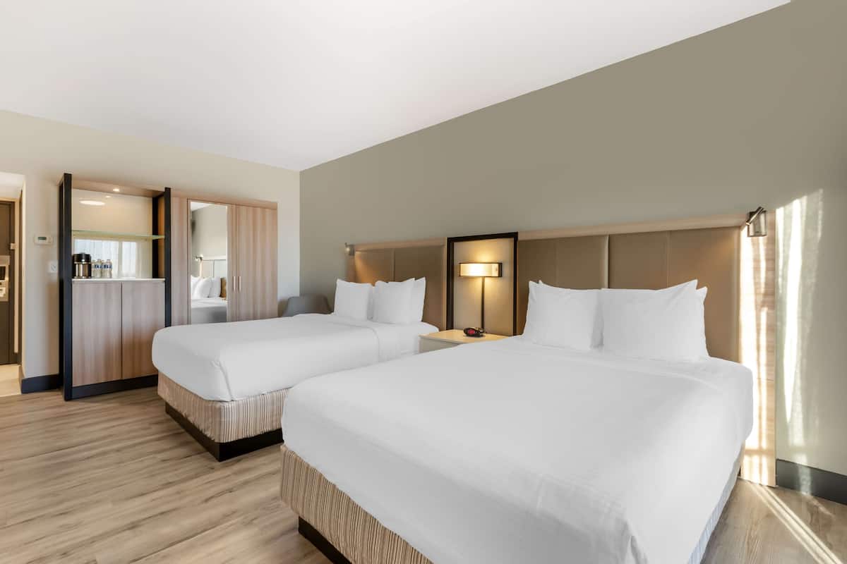 A hotel room with two beds, a bedside table with a lamp, a wardrobe, and a bathroom visible through an open door.