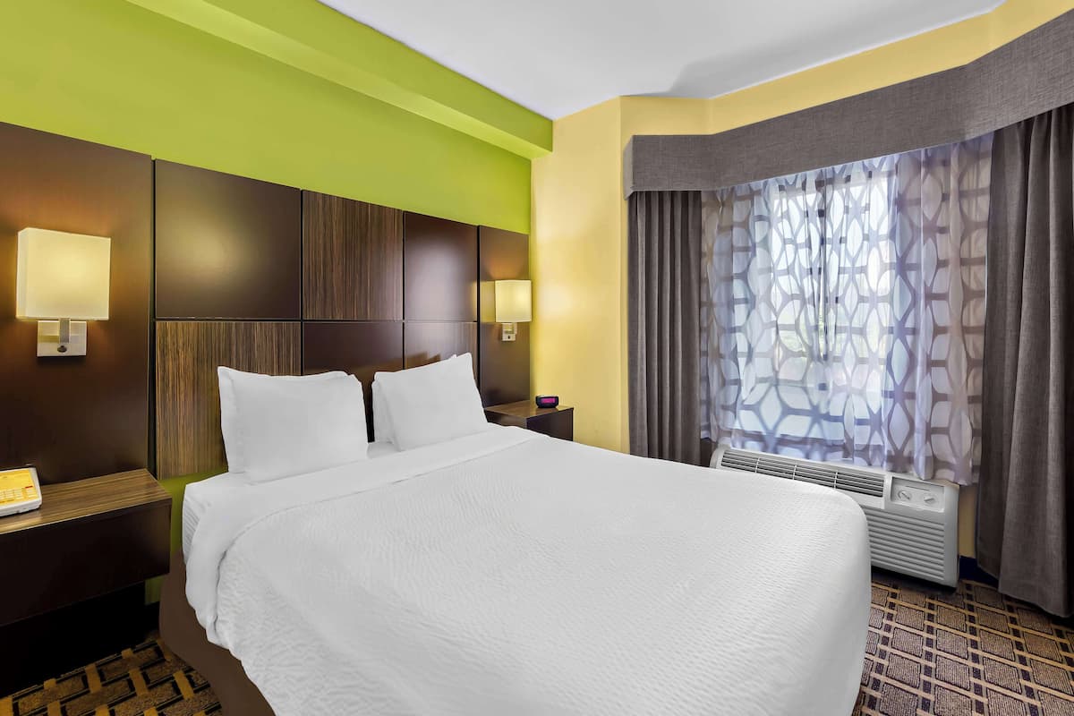 A hotel room with a large bed, two bedside lamps, a nightstand, a heater, and a window with patterned curtains.