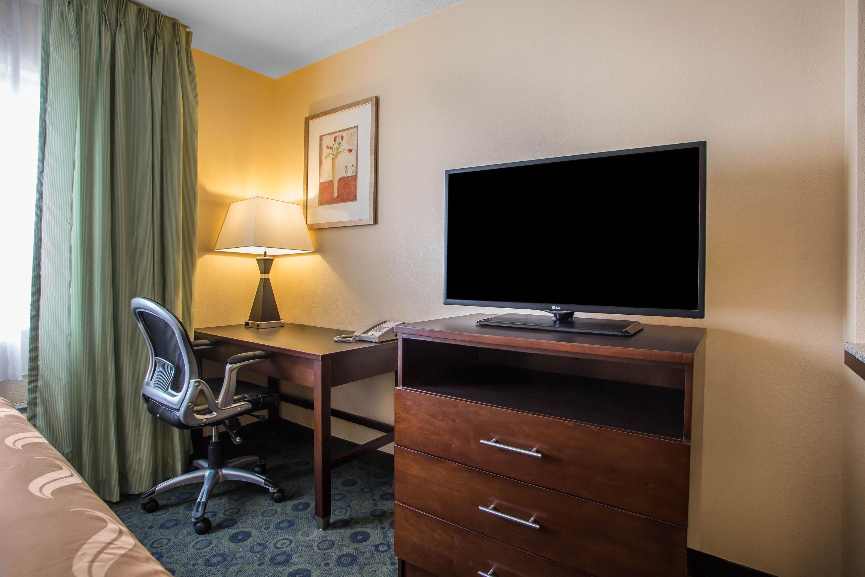 A hotel room with a desk, chair, TV, lamp, and a bed.