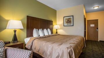 A hotel room with a large bed, two bedside lamps, a nightstand with a lamp, a chair, and a door.