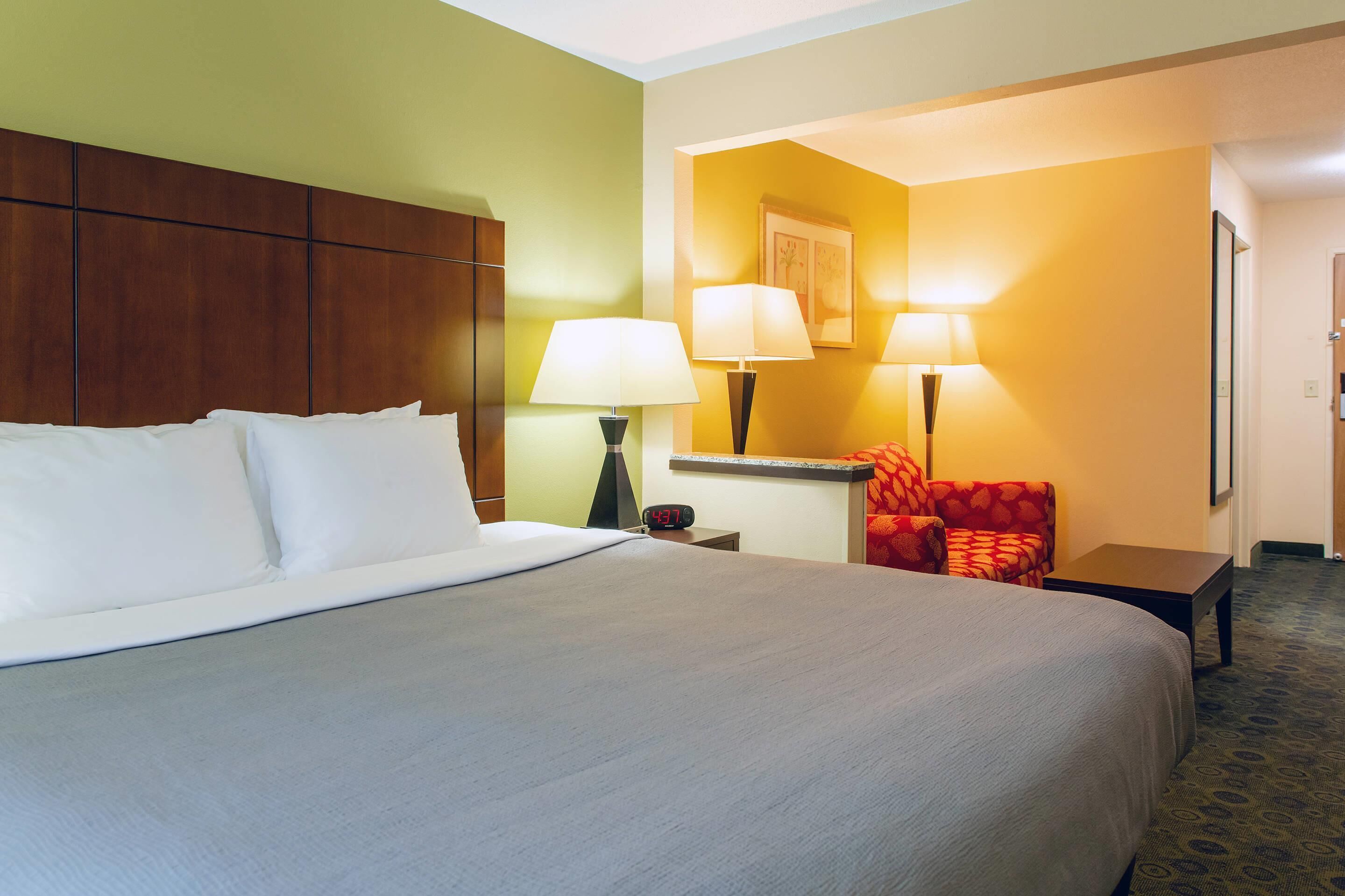 A hotel room with a large bed, two bedside lamps, a red armchair, a small table, and a nightstand.
