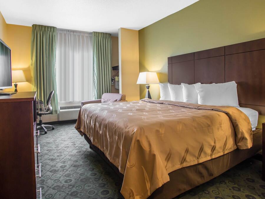 A hotel room with a large bed, two bedside tables, a desk with a computer, a chair, and a window with curtains.